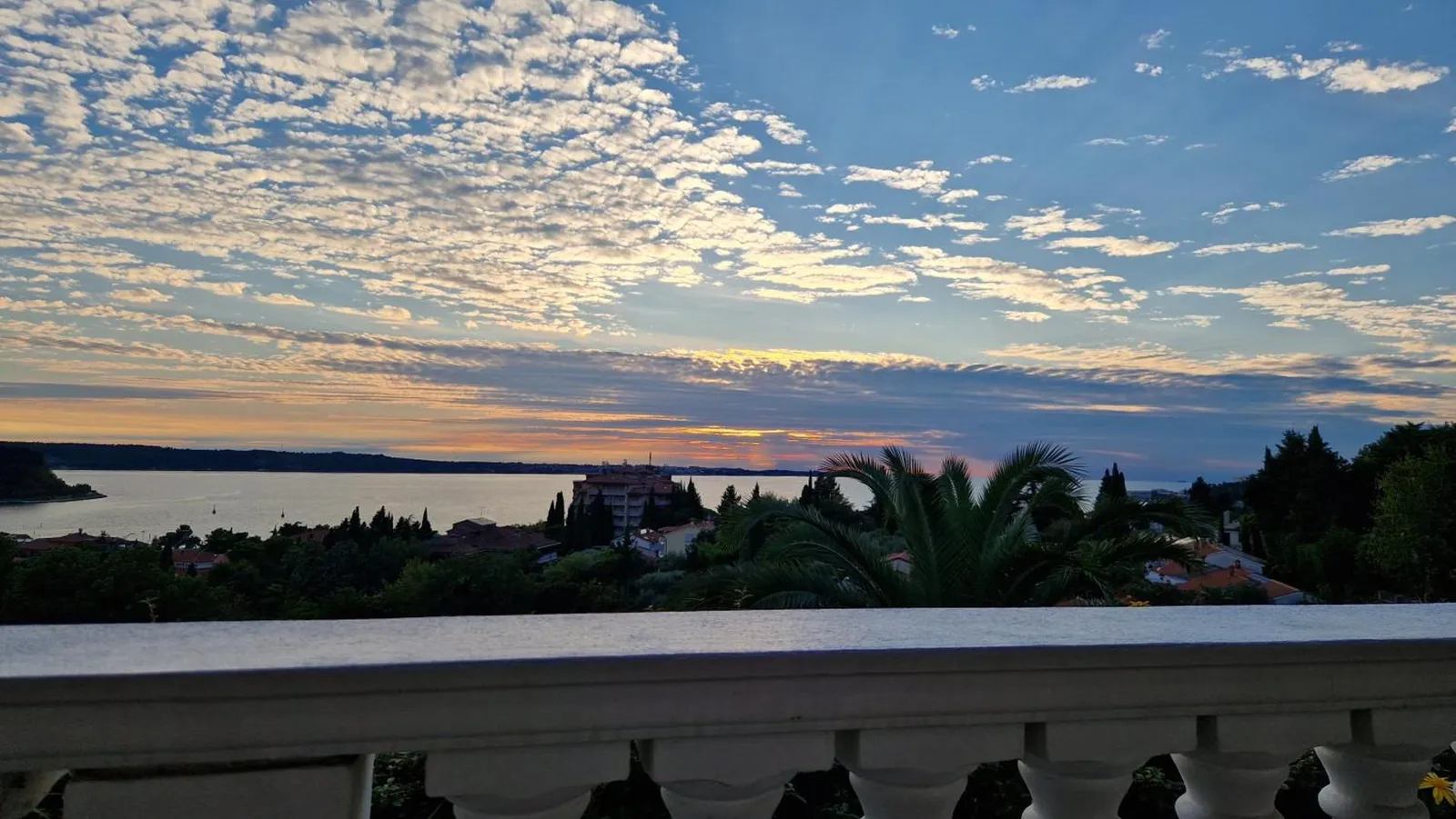 Sončni zahod nad Portoroškim zalivom z balkona vile, spredaj bela balustrada in palme
